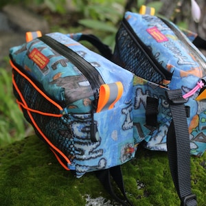 May include: Two blue and black patterned bike bags with orange straps and black mesh panels. The bags have a zipper closure and are attached to each other with a black strap.
