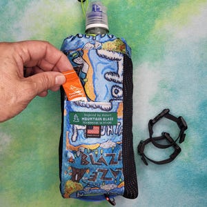 May include: A blue water bottle holder with a colorful mountain and campfire print, featuring a black mesh side pocket, a green drawstring cord, and a label reading Inspired by Nature MOUNTAIN BLAZE HANDMADE IN MAINE with a small US flag.