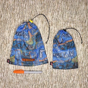 May include: Two blue mesh drawstring bags with colourful graphics and the word "MOUNTAIN" in white. A small orange and grey marker is placed below the bags. The bags have black drawstrings and yellow and black cord locks.