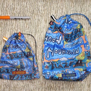 May include: Two blue drawstring bags with a mountain-themed design, featuring the words "Mountain Blaze." The bags have a colorful pattern with orange, green, and white accents. An orange Sharpie pen is also visible.