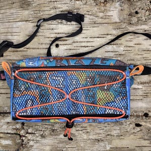 May include: A rectangular waist bag featuring a blue fabric base with a colourful graphic print, a black mesh front pocket, and bright orange bungee cord detailing. The bag includes an adjustable black strap with a plastic buckle and side tabs.