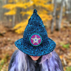 May include: A blue and black knit witch hat with a pointed top and wide brim. The hat features a circular patch with a white star design, outlined in yellow and red, on a green background. The hat is worn by a person with purple hair.