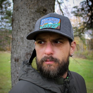 Peut inclure: Casquette de baseball grise à chevrons avec un écusson "Mountain Blaze" en bleu, vert et blanc. La casquette a une visière incurvée et est portée à l'extérieur.