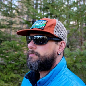 May include: A burnt orange and tan mesh-backed cap with a mountain scene patch. The cap has a dark brown brim and is paired with black sunglasses. The person is wearing a blue jacket.
