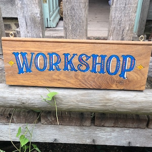 May include: A wooden sign displaying the word "WORKSHOP" in blue, decorative lettering. The sign is rectangular with hanging loops. The background features a wooden bench, greenery, and a partially visible door.