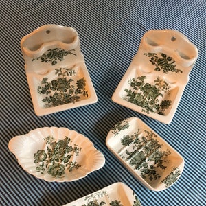 May include: Set of five vintage ceramic soap dishes with a green floral pattern. The dishes are in various shapes and sizes, including a wall-mounted soap dish with a toothbrush holder.