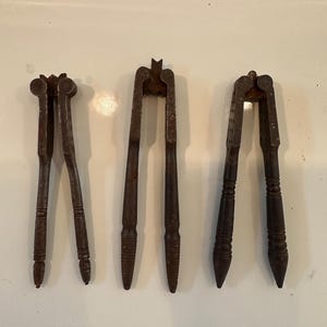 May include: Three dark brown metal nutcrackers arranged vertically. Each tool features a hinged mechanism at the top with a textured gripping area and long, slender handles that taper to a point at the bottom, showing signs of age and wear.