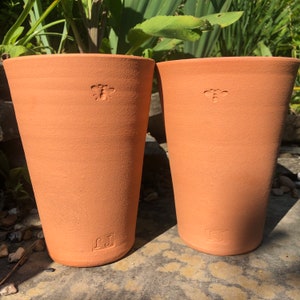 May include: Two terracotta plant pots. Each pot features a bee emblem near the top and the initials "JJ" near the base. The pots are cylindrical with a gently flared rim. The unglazed pots have a textured surface and are a warm, earthy colour.