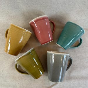 May include: Five ceramic mugs in mustard yellow, dusty rose, teal, olive green, and grey. Each mug has a white rim and a curved handle. The mugs are arranged on a neutral-coloured surface.