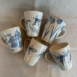 May include: A set of three ceramic mugs with a speckled, matte finish. The mugs are in shades of cream, light grey, and dark grey. Each mug has a simple, minimalist design, ideal for coffee or tea.
