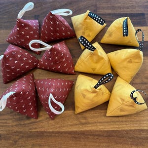 May include: A collection of ten pyramid-shaped fabric bean bags. Four are dark red with a small white floral pattern and white ribbon loops. Six are mustard yellow with a subtle geometric triangle pattern and black and white chequered ribbon loops.