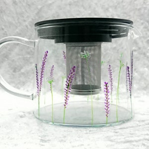 Puede incluir: Una tetera de vidrio transparente con un dise&ntilde;o floral de lavanda. La tetera tiene un infusor de acero inoxidable extra&iacute;ble y una tapa negra.