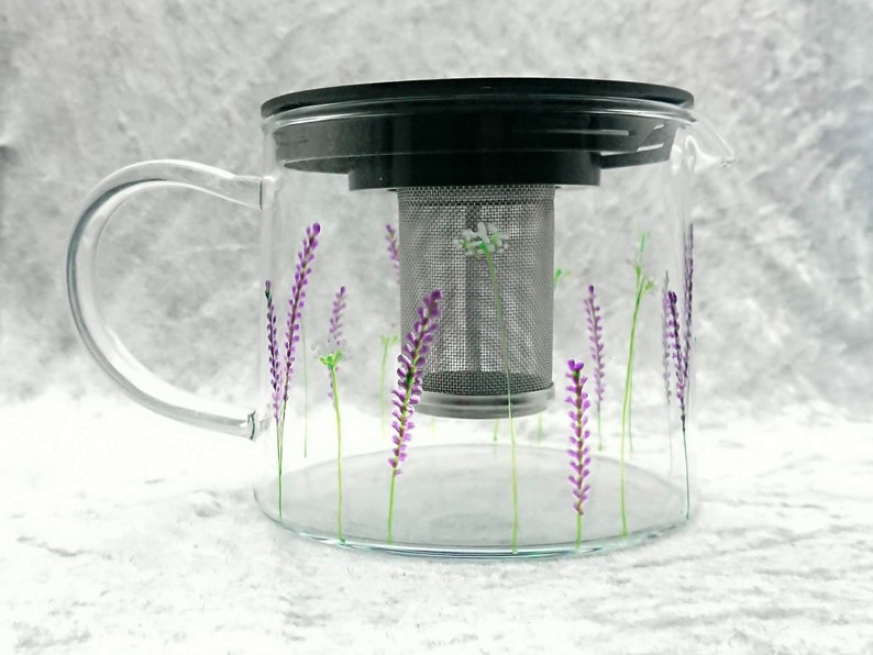 Puede incluir: Una tetera de vidrio transparente con un dise&ntilde;o floral de lavanda. La tetera tiene un infusor de acero inoxidable extra&iacute;ble y una tapa negra.