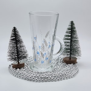 May include: A clear glass mug with a handle, hand-painted with a white and blue floral design. The mug is surrounded by silver beads and two small artificial pine trees.