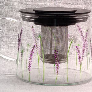 Puede incluir: Una tetera de vidrio con tapa negra y un filtro de acero inoxidable. La tetera est&aacute; decorada con flores de lavanda pintadas a mano y tallos verdes.