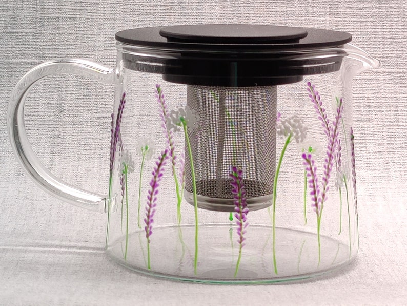 Puede incluir: Una tetera de vidrio con tapa negra y un filtro de acero inoxidable. La tetera est&aacute; decorada con flores de lavanda pintadas a mano y tallos verdes.