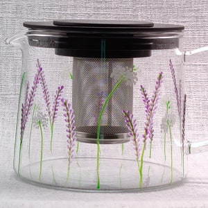 Puede incluir: Una tetera de vidrio transparente con un filtro de acero inoxidable y una tapa negra. La tetera est&aacute; decorada con un dise&ntilde;o floral de flores de lavanda moradas y flores blancas.