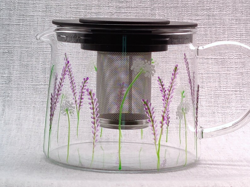 Puede incluir: Una tetera de vidrio transparente con un filtro de acero inoxidable y una tapa negra. La tetera est&aacute; decorada con un dise&ntilde;o floral de flores de lavanda moradas y flores blancas.