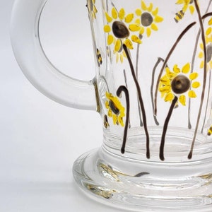 May include: A clear glass mug with a handle, decorated with hand-painted sunflowers and bees. The sunflowers are yellow with brown centres, and the bees are black and yellow.