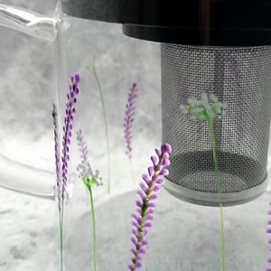 Puede incluir: Una taza de t&eacute; de vidrio transparente con un colador incorporado. La taza tiene un dise&ntilde;o floral morado y blanco.