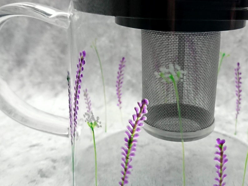 Puede incluir: Una taza de t&eacute; de vidrio transparente con un colador incorporado. La taza tiene un dise&ntilde;o floral morado y blanco.
