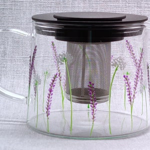 Puede incluir: Una tetera de vidrio transparente con tapa negra y un infusor de acero inoxidable. La tetera est&aacute; decorada con flores de lavanda pintadas a mano.