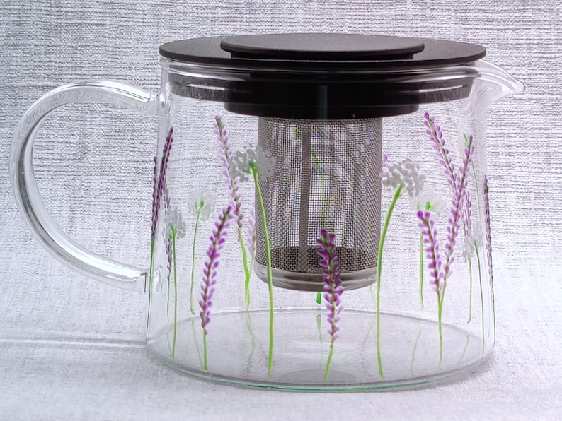 Puede incluir: Una tetera de vidrio transparente con tapa negra y un infusor de acero inoxidable. La tetera est&aacute; decorada con flores de lavanda pintadas a mano.