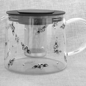 May include: A clear glass teapot with a black lid and a stainless steel infuser. The teapot is decorated with black roses and white dots.