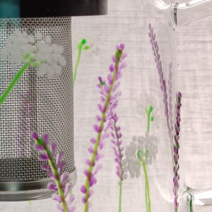 Puede incluir: Una jarra de vidrio transparente con un filtro de malla de acero inoxidable y un dise&ntilde;o floral. El dise&ntilde;o presenta flores moradas y blancas.