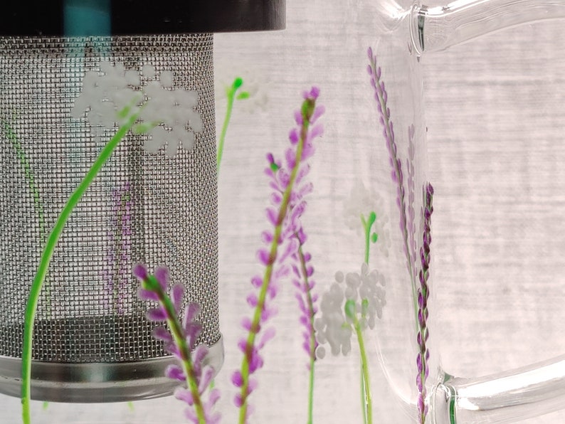 Puede incluir: Una jarra de vidrio transparente con un filtro de malla de acero inoxidable y un dise&ntilde;o floral. El dise&ntilde;o presenta flores moradas y blancas.