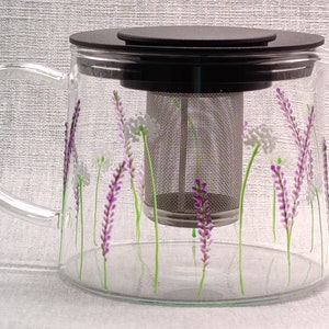 Puede incluir: Una tetera de vidrio con tapa negra y un filtro de acero inoxidable. La tetera est&aacute; decorada con flores de lavanda pintadas a mano y tallos verdes.