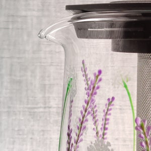 Puede incluir: Una tetera de vidrio transparente con una tapa negra y un filtro de malla de acero inoxidable. La tetera tiene un dise&ntilde;o floral con flores moradas y blancas.
