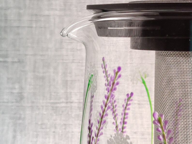 Puede incluir: Una tetera de vidrio transparente con una tapa negra y un filtro de malla de acero inoxidable. La tetera tiene un dise&ntilde;o floral con flores moradas y blancas.