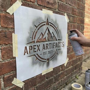 May include: A white rectangular stencil featuring a mountain range graphic inside a compass rose design with the text APEX ARTIFACTS and EST. 2023. The stencil is taped to a brick wall, and a spray paint can is being used to apply paint.