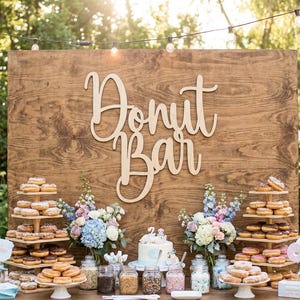 May include: A wooden sign that reads "Donut Bar" is displayed above a table laden with donuts, a cake, and various toppings. The donuts are arranged on tiered wooden stands and white pedestals, with floral arrangements adding color.
