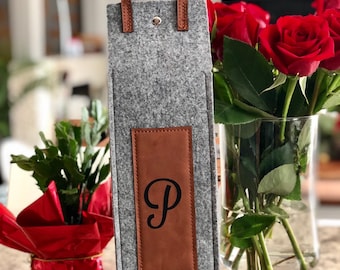 May include: A gray felt wine bottle carrier with brown leather handles and a brown leather patch with the letter "P" in black. The carrier is sitting on a table next to a vase of red roses.
