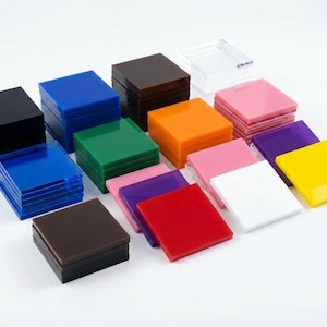 May include: A collection of colorful square acrylic blocks in various shades, including black, blue, green, orange, pink, purple, red, white, and yellow. The blocks are stacked and arranged on a white surface.