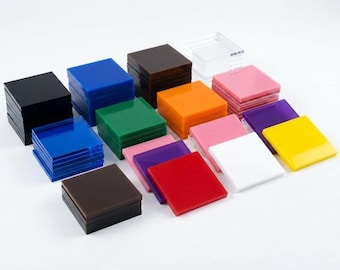 Puede incluir: Colección de bloques cuadrados de acrílico de colores en varios tonos, incluyendo negro, azul, verde, naranja, rosa, morado, rojo, blanco y amarillo. Los bloques están apilados sobre una superficie blanca.