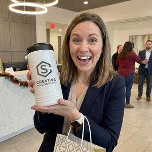 May include: A white tumbler with a black lid, featuring the logo and text "CREATIVE SOLUTIONS, INC." is held by a smiling person. The background includes a decorated reception desk and other people in an office setting.