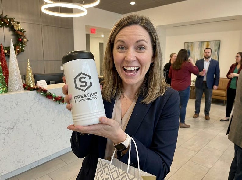 May include: A white tumbler with a black lid, featuring the logo and text "CREATIVE SOLUTIONS, INC." is held by a smiling person. The background includes a decorated reception desk and other people in an office setting.