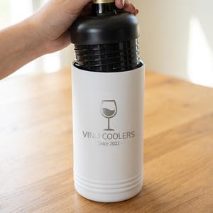 May include: A white wine cooler with a black top, holding a wine bottle. The cooler has the text "Vino Coolers Since 2023" and a wine glass graphic. The cooler is cylindrical and sits on a wooden surface.