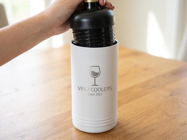 May include: A white wine cooler with a black top, holding a wine bottle. The cooler has the text "Vino Coolers Since 2023" and a wine glass graphic. The cooler is cylindrical and sits on a wooden surface.