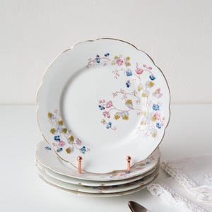 Op de afbeelding: Een stapel witte porseleinen borden met een geschulpte rand en gouden rand. Elk bord is versierd met een bloemenpatroon in roze, blauw en groen. De borden worden gepresenteerd op een koperkleurige bordenstandaard.