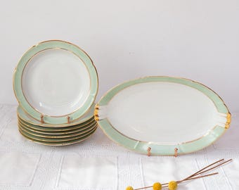 May include: A set of vintage white porcelain plates and a serving platter with a mint green and gold trim. The plates are stacked, and the platter is oval-shaped. The set is displayed on a white tablecloth.