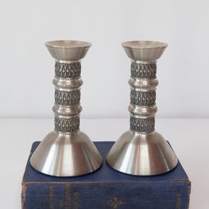 Op de afbeelding: Twee zilverkleurige metalen kandelaars met een decoratief ruitpatroon op de centrale kolom. De kandelaars hebben een uitlopende basis en een platte bovenkant. Ze staan op een blauw boek.