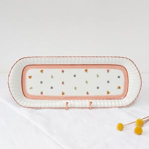 May include: A rectangular ceramic serving tray with a white base, pink border, and floral design. The tray has a scalloped edge and is supported by a copper-coloured stand. The floral pattern features small yellow, pink, and blue flowers.