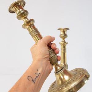 Può includere: Due candelabri in ottone ornati. Il candelabro principale è tenuto ad angolo, mostrando il suo design dettagliato. La base e lo stelo presentano motivi intricati. Un secondo candelabro è visibile sullo sfondo. La parola "Beauté" è tatuata sul braccio.