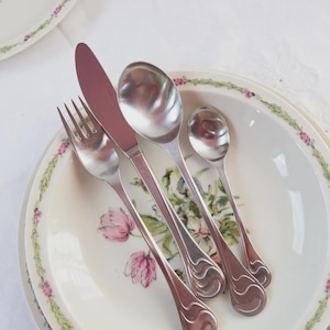 Puede incluir: Un juego de cubiertos de acero inoxidable, que incluye un tenedor, un cuchillo y cucharas, dispuestos sobre un plato decorado con motivos florales. El plato tiene un fondo blanco con diseños florales rosas y verdes. La cubertería tiene un diseño de mango decorativo.