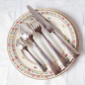 May include: A collection of vintage silver cutlery, comprising a knife, fork, and two spoons, presented on a floral patterned plate with a gold rim. The plate rests on a white lace tablecloth. The cutlery is silver.