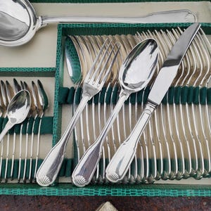 Peut inclure: Un ensemble de couverts en argent poli, comprenant des fourchettes, des cuillères et des couteaux, disposés dans un étui doublé de vert. Une grande louche est également visible. La coutellerie présente un design simple et élégant.
